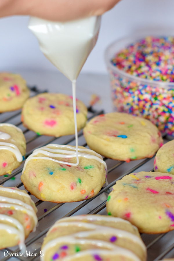 Sprinkle cookies on cooling rack with a side of sprinkles next to them. A piping bag filled with icing that is drizzled over cookies.