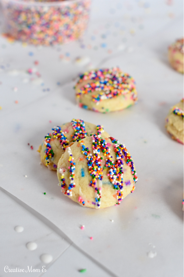 Birthday cake cookies on parchment paper with sprinkles and icing scattered on the side of it. With a bowl of sprinkles in the background.
