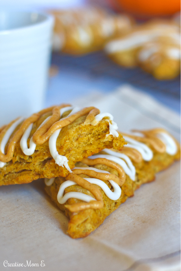 Classic pumpkin spice scones on a napkin with a bite taken out of one with a coffee and more fluffy pumpkin scones in the background. 
