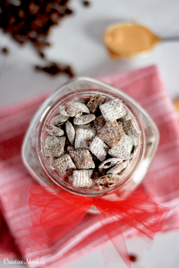 Peanut Butter Muddy Buddies in a clear jar sitting on a red tea towel with  chocolate chips and a scoop of peanut butter on the side. 