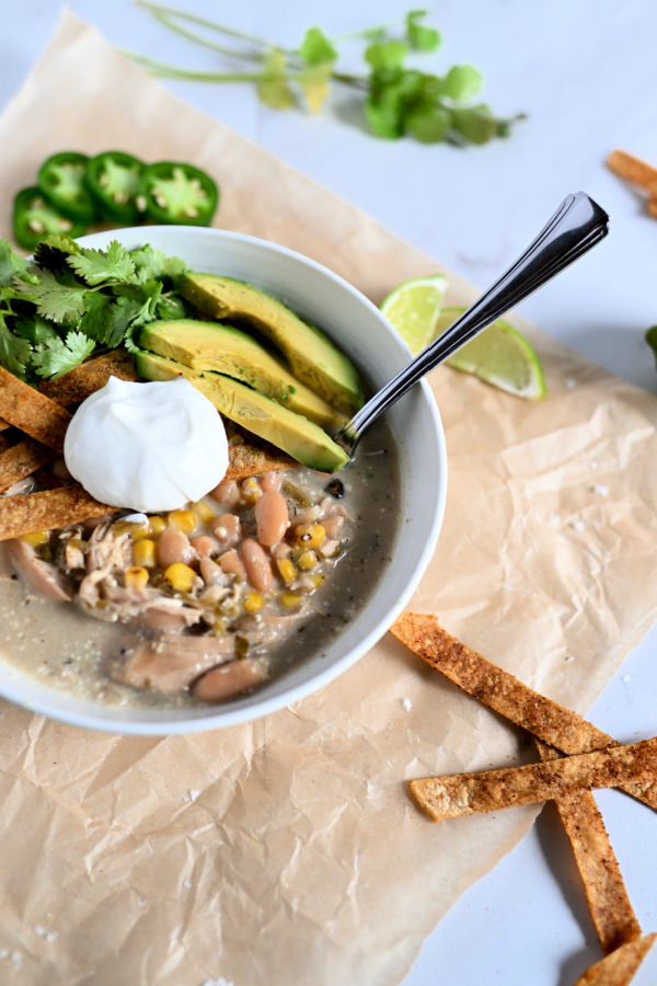 White bowl of chicken chili with sour cream, avocado, cilantro, on a brown paper with tortilla strips and lime.