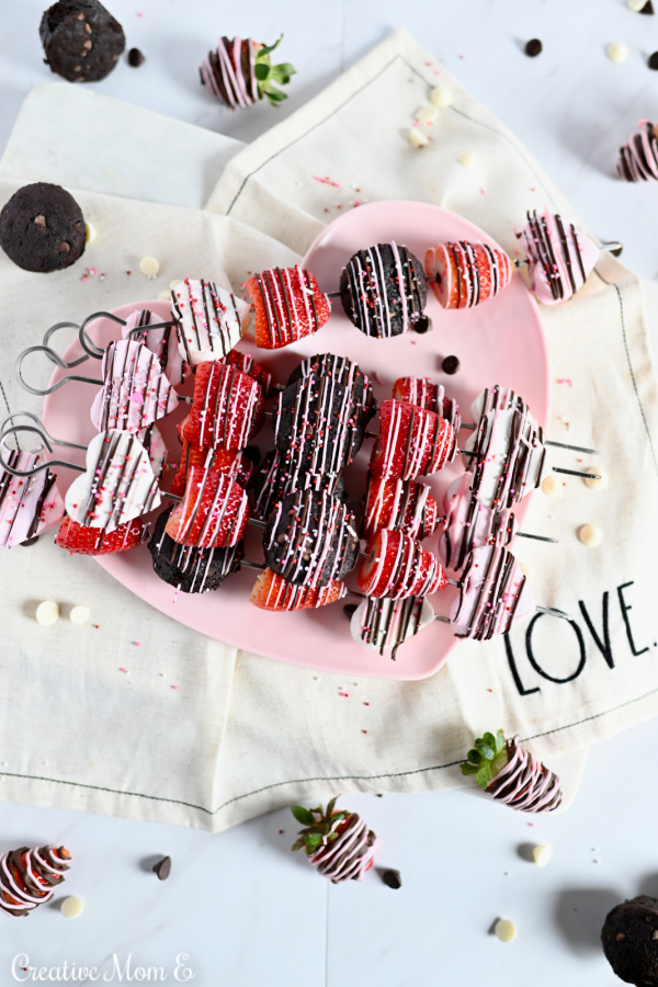 Strawberry, marshmallow and brownie on a skewer drizzled with chocolate and sprinkles on a pink heart plate.