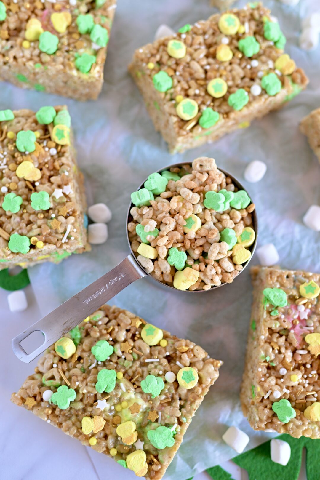 Lucky Charms Marshmallow Rice Krispies squares on parchment paper with a measuring cup filled with lucky charm cereal.