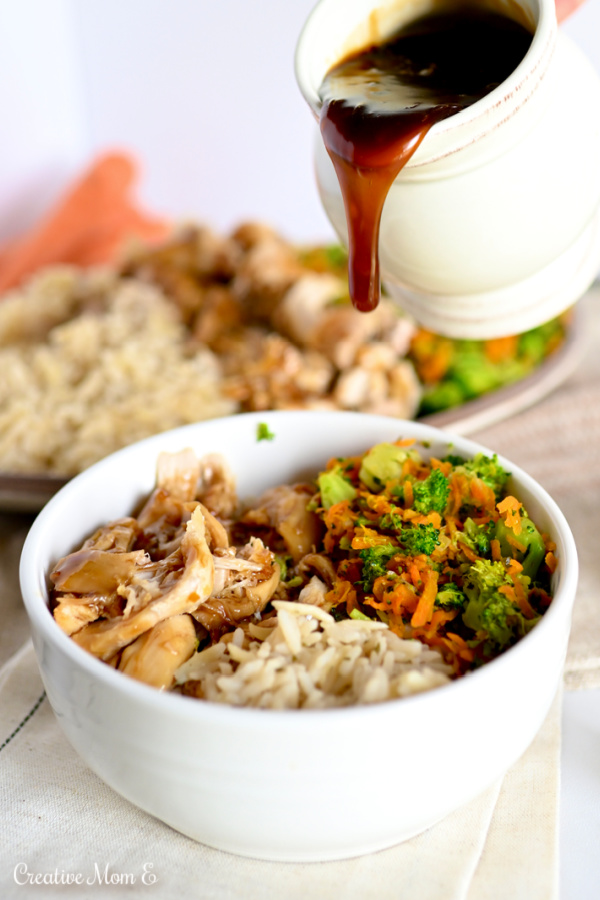 A white bowl filled with chicken, vegetables and rice. A small pitcher of chicken teriyaki drizzling over it.
