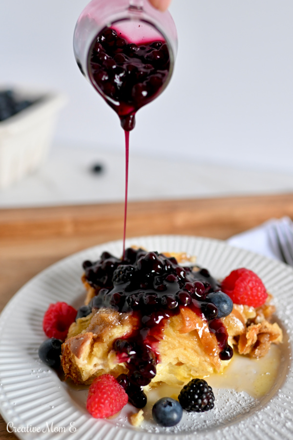 Homemade blueberry syrup being drizzled over cream cheese French toast casserole with fresh berries. 