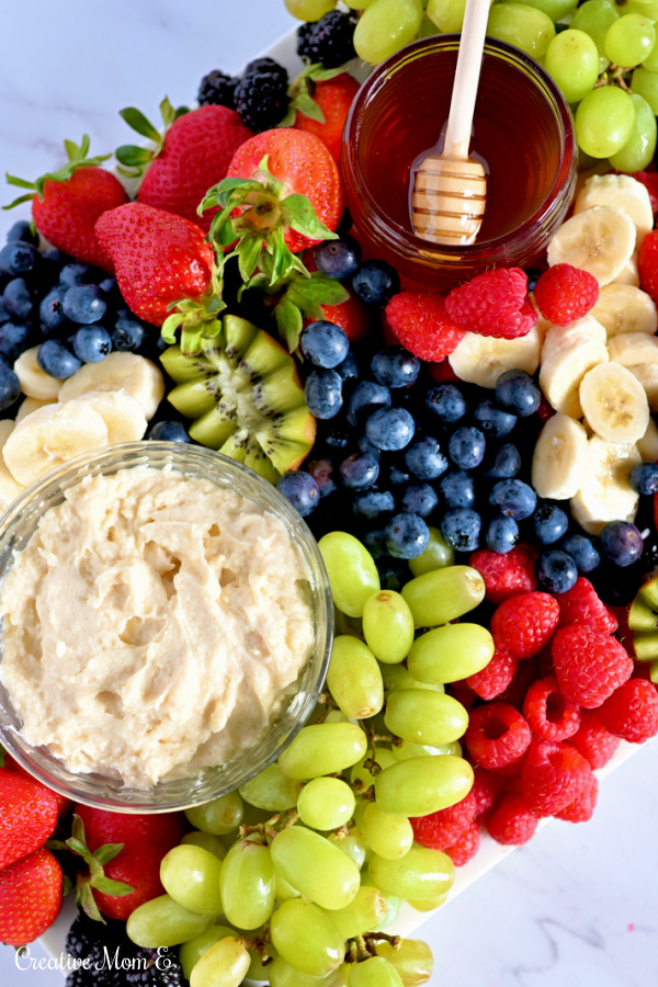 Fruit board filled with berries, bananas, kiwi and grapes next to fruit dip and honey. 
