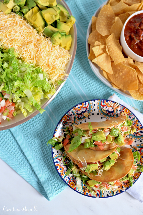 Taco night spread with prepared tacos on a plate next to a platter filled with tomato, lettuce, cheese and avocados. Enjoy with chips and salsa.